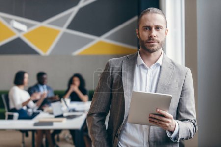 Téléchargez les photos : Un jeune homme d'affaires confiant tenant une tablette numérique pendant que ses collègues travaillent sur fond - en image libre de droit