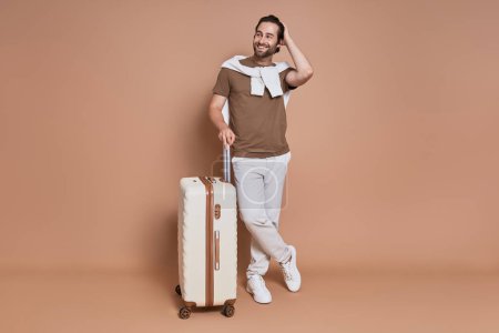 Téléchargez les photos : Pleine longueur de joyeux jeune homme avec valise debout sur fond brun - en image libre de droit