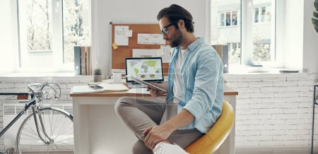 Téléchargez les photos : Beau jeune homme utilisant une tablette numérique tout en étant assis à son lieu de travail - en image libre de droit