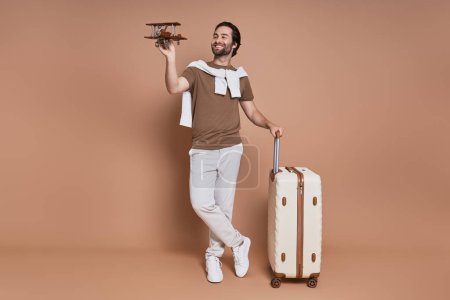 Téléchargez les photos : Pleine longueur de jeune homme heureux avec valise tenant l'avion en bois sur fond brun - en image libre de droit
