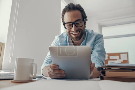 Téléchargez les photos : Beau jeune homme travaillant sur tablette numérique et souriant tout en étant assis à son lieu de travail au bureau - en image libre de droit