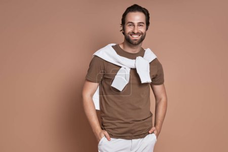 Téléchargez les photos : Jeune homme à la mode tenant la main dans les poches et souriant sur fond brun - en image libre de droit