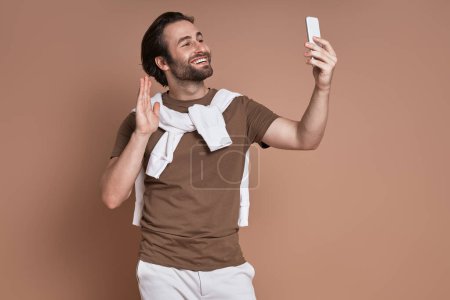 Foto de Joven feliz mirando su teléfono inteligente y agitando la mano contra el fondo marrón - Imagen libre de derechos