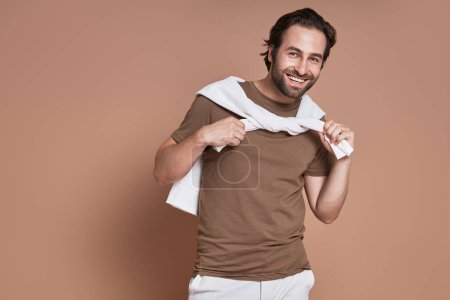 Téléchargez les photos : Jeune homme à la mode regardant la caméra et souriant sur fond brun - en image libre de droit