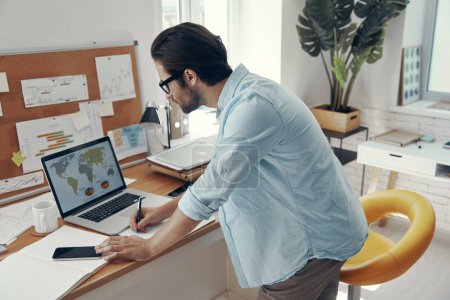 Téléchargez les photos : Jeune homme concentré utilisant un ordinateur portable et prenant des notes tout en se tenant près de son lieu de travail - en image libre de droit