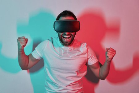 Téléchargez les photos : Jeune homme excité en réalité virtuelle lunettes geste avec des ombres colorées en arrière-plan - en image libre de droit