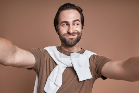 Téléchargez les photos : Jeune homme ludique faisant selfie et grimaçant tout en se tenant debout sur fond brun - en image libre de droit