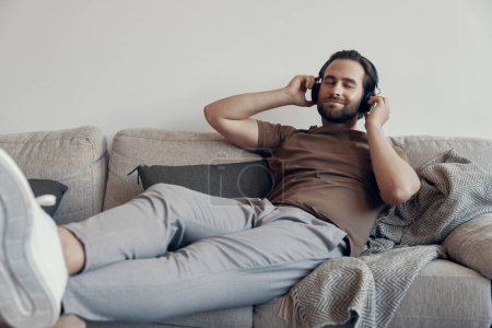 Foto de Hombre joven guapo ajustando sus auriculares mientras se relaja en el sofá en casa - Imagen libre de derechos