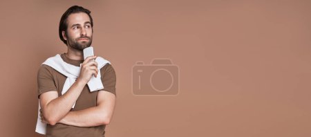 Foto de Hombre joven guapo sosteniendo el teléfono inteligente y mirando reflexivo mientras está de pie contra el fondo marrón - Imagen libre de derechos