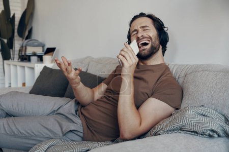 Téléchargez les photos : Joyeux jeune homme dans les écouteurs en utilisant un téléphone intelligent comme un microphone tout en se relaxant sur le canapé - en image libre de droit