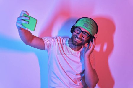 Photo for Happy young man in headphones making selfie by smart phone against colorful background - Royalty Free Image
