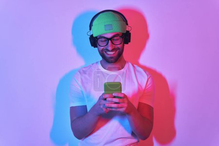 Foto de Hombre joven guapo en auriculares con teléfono inteligente y sonriendo contra el fondo colorido - Imagen libre de derechos