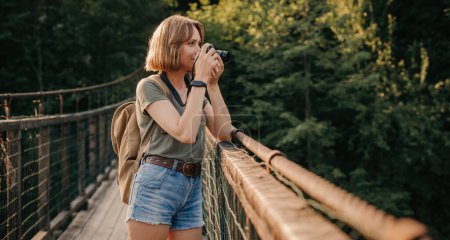 Foto de Atractiva joven viajera fotografiando con cámara mientras está de pie en el puente escénico - Imagen libre de derechos