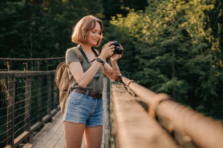 Foto de Sonriente joven viajera fotografiando con la cámara mientras está de pie en el puente escénico - Imagen libre de derechos
