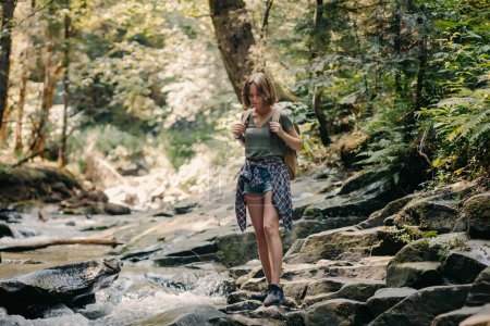 Foto de Longitud completa de la joven viajera con mochila caminando por las piedras a lo largo del río de montaña - Imagen libre de derechos