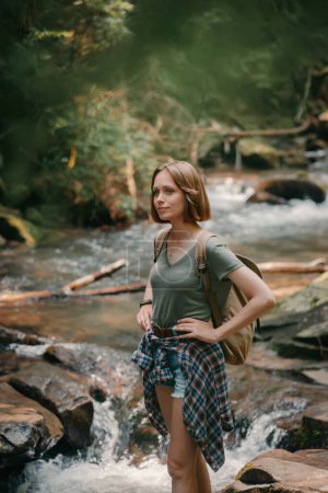 Téléchargez les photos : Jeune voyageuse confiante avec sac à dos debout au bord de la rivière de montagne et bénéficiant d'une vue - en image libre de droit