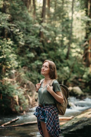 Foto de Joven viajera sonriente con mochila junto al río de montaña y disfrutando de las vistas - Imagen libre de derechos
