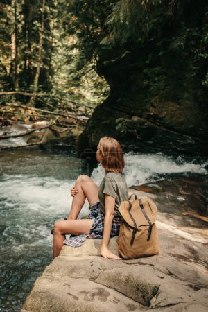 Foto de Joven viajera con mochila descansando sentada junto al río de la montaña - Imagen libre de derechos