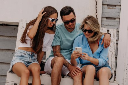 Foto de Tres jóvenes amigos mirando el teléfono inteligente mientras están sentados en la paleta de madera al aire libre - Imagen libre de derechos