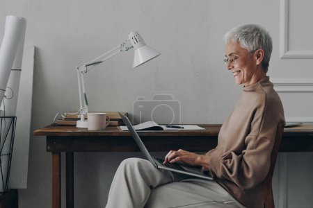 Foto de Elegante mujer de negocios senior usando laptop y sonriendo mientras está sentada en su lugar de trabajo - Imagen libre de derechos