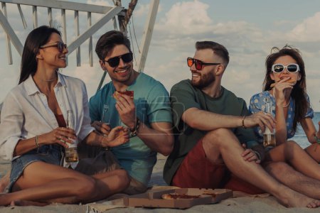 Foto de Grupo de jóvenes felices disfrutando de pizza y cerveza mientras pasan tiempo juntos en la playa - Imagen libre de derechos