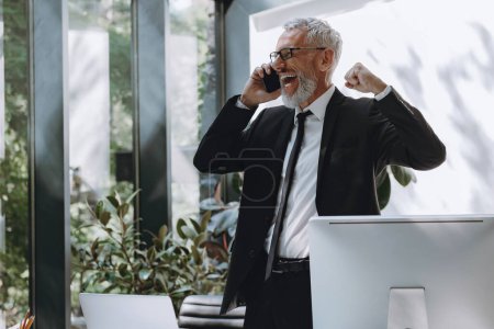 Foto de Feliz hombre de negocios maduro hablando por teléfono móvil y haciendo gestos mientras trabaja en la oficina - Imagen libre de derechos