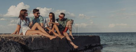 Foto de Grupo de jóvenes amigos felices disfrutando de la cerveza mientras pasan tiempo despreocupados junto al mar juntos - Imagen libre de derechos