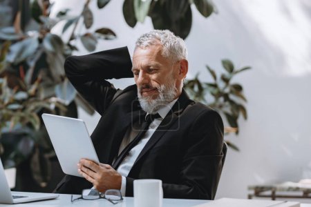 Foto de Hombre maduro ocupado en ropa formal usando tableta digital mientras trabaja en la oficina moderna - Imagen libre de derechos