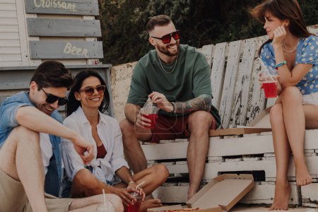 Foto de Grupo de amigos felices disfrutando de pizza y cócteles mientras se relajan cerca de la cafetería de la playa juntos - Imagen libre de derechos
