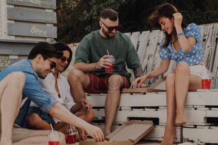 Foto de Grupo de amigos felices disfrutando de comida y bebidas mientras se relajan cerca de la cafetería de la playa juntos - Imagen libre de derechos