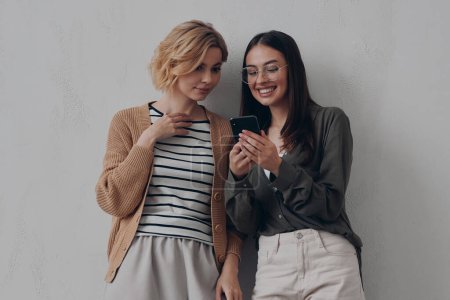 Foto de Dos hermosas mujeres jóvenes usando el teléfono inteligente mientras están de pie cerca de su lugar de trabajo creativo - Imagen libre de derechos