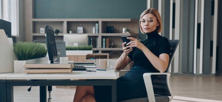 Foto de Mujer de negocios de confianza sosteniendo el teléfono inteligente y mirando a la cámara mientras está sentado en la oficina - Imagen libre de derechos
