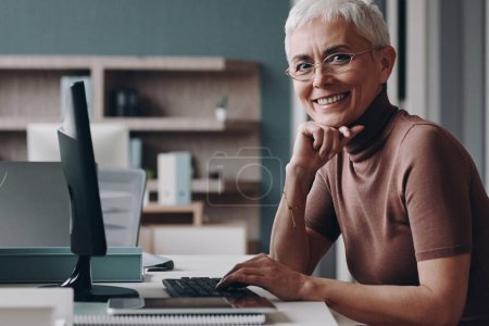 Foto de Mujer de negocios mayor mirando a la cámara y sonriendo mientras está sentada en su lugar de trabajo en la oficina - Imagen libre de derechos