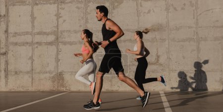 Foto de Tres jóvenes confiados en ropa deportiva corriendo frente a la pared de hormigón al aire libre juntos - Imagen libre de derechos