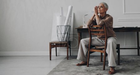 Foto de Hermosa mujer mayor mirando reflexivo mientras se sienta en su lugar de trabajo creativo - Imagen libre de derechos