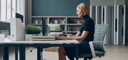 Foto de Joven empresaria seria usando el ordenador portátil mientras está sentada en su lugar de trabajo en la oficina - Imagen libre de derechos