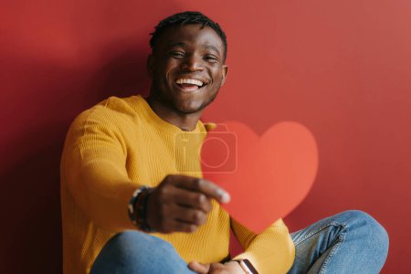 Foto de Joven hombre africano alegre mostrando tarjeta de San Valentín en forma de corazón mientras está sentado sobre fondo rojo - Imagen libre de derechos