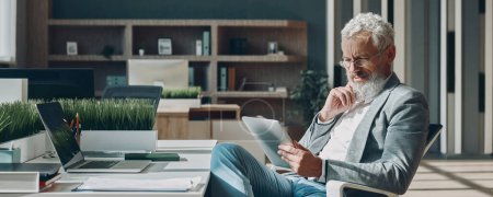 Foto de Confiado hombre de negocios maduro utilizando tableta digital mientras está sentado en su lugar de trabajo en la oficina - Imagen libre de derechos