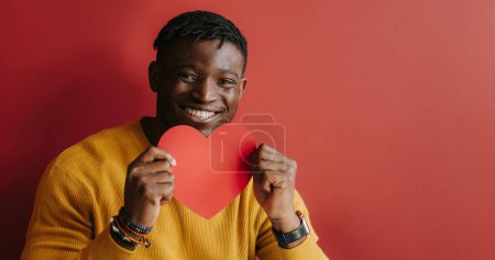 Foto de Feliz joven africano sosteniendo la tarjeta de San Valentín en forma de corazón mientras está sentado sobre fondo rojo - Imagen libre de derechos