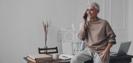 Foto de Mujer de negocios mayor hablando por teléfono y sonriendo mientras está sentada en el escritorio en la oficina - Imagen libre de derechos