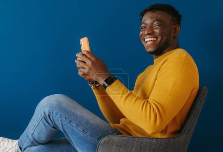 Foto de Joven hombre africano alegre en ropa casual usando un teléfono inteligente mientras está sentado en la silla contra la pared azul - Imagen libre de derechos