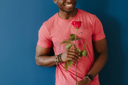 Foto de Encantador joven africano en ropa casual sosteniendo rosa soltera y sonriendo sobre fondo azul - Imagen libre de derechos