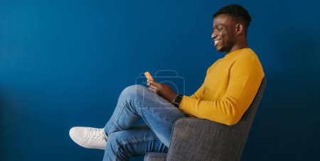 Foto de Feliz joven africano en ropa casual usando un teléfono inteligente mientras está sentado en la silla contra la pared azul - Imagen libre de derechos