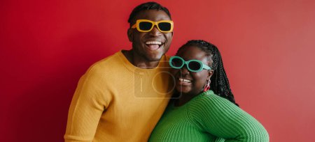 Téléchargez les photos : Jeune couple africain à la mode en lunettes tendance embrassant et souriant sur fond rouge ensemble - en image libre de droit