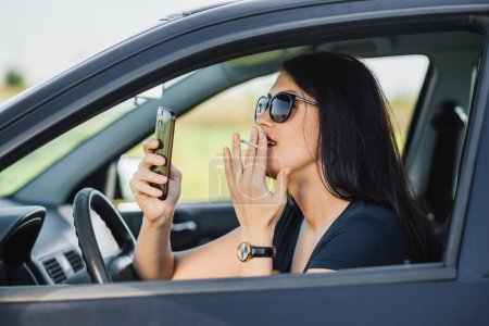 Photo for Young woman smoking in the car and looking on the phone while the car is stopped - Royalty Free Image