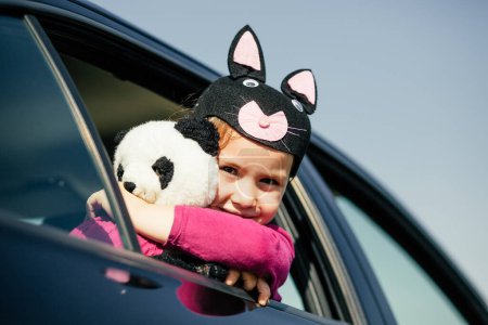 Photo for Beautiful little girl wearing a funny hat and holding a teddy bear in her arms while looking to the camera between car's window - Royalty Free Image
