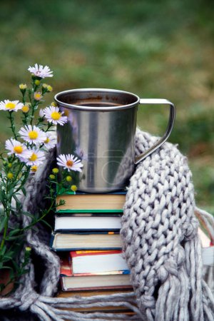 Téléchargez les photos : Une tasse de café sur un fond de livres et une couverture. Composition d'une tasse de café, de fleurs, de livres et d'une couverture dans la nature. - en image libre de droit