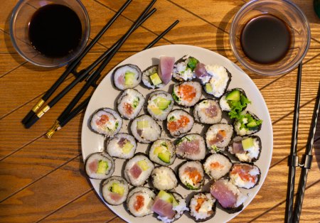 Ein mit verschiedenen Sushi-Rollen gefüllter Teller neben einem Paar Essstäbchen auf einem Holztisch.