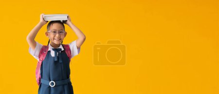 Foto de Back to school banner idea concept, Happy asian school girl in uniform  hold books on her head, isolated on yellow background with Clipping paths for design work empty free space - Imagen libre de derechos