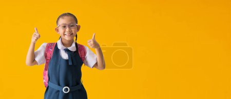 Foto de Back to school banner idea concept, Happy asian school girl in uniform with show thumbs up for good, isolated on yellow background with Clipping paths for design work empty free space - Imagen libre de derechos